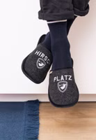 baumelnd ein Paar schwarze Filzpantoffel mit weißer Stickerei eines röhrenden Hirsches im Wappen sowie dem Schriftzug Platzhirsch