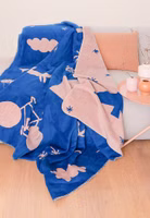 eine rosa und dunkelblaue Kuscheldecke mit abstrakten Motiven von Blumen, Fahrrad, Katze, Mond liegend auf einem Sofa im Wohnzimmer