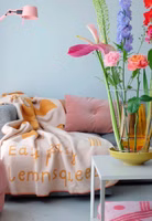 eine senfgelb- und beigefarbene Easy Peasy Kuscheldecke mit Zitronenmotiv und Schriftzug liegend auf einer Couch und bunte Blumen im Vordergrund von PRCHTG