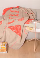 eine beige- und korallefarbene Kuscheldecke mit abstrakten Häusern sowie Schriftzug Home sweet Home liegend auf einer hellgrauen Couch
