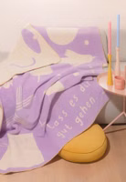 eine creme- und lilafarbene Kuscheldecke mit abstrakten Motiven und Lass es dir gut gehen Schriftzug liegend auf einer Couch im Wohnzimmer