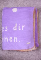 eine gefaltete creme- und lilafarbene Kuscheldecke mit abstrakten Motiven und Lass es dir gut gehen Schriftzug und gelber Umrandung