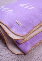 eine gefaltete creme- und lilafarbene Kuscheldecke mit abstrakten Motiven und Lass es dir gut gehen Schriftzug und gelber Umrandung