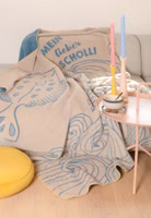 eine beige Kuscheldecke mit dem blaugrauen Motiv einer Scholle und den schwungvollen Konturen von Meereswellen sowie dem Schriftzug Mein lieber Scholli liegend auf einer Couch im Wohnzimmer