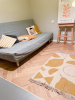Adelheid X Anna Jansen Easy Peasy Teppich 80 x 120 cm in einem Zimmer vor einem Schlafsofa liegend ein beige senffarbener "Easy peasy" Teppich mit abstraktem Zitronenmotiv