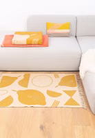 Teppich Easy Peasy von Adelheid X Anna Katharina Jansen in einem Wohnzimmer vor einer hellgrauen Couch liegend ein beige senffarbener "Easy peasy" Teppich mit abstraktem Zitronenmotiv