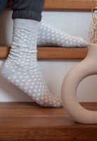 jemand sitzend auf Treppenstufen mit einem Paar hellgraue Glücksbringer Socken mit weißen Punkten, uni grau abgesetztem Bündchen-, Fersen- und Zehenbereich sowie oberhalb des Knöchels dem Motiv einer Edelweiß Blüte und dem Schriftzug Glücksbringer