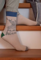 sitzend auf Treppenstufen mit einem Paar graubeigen Platzhirsch Socken mit grauem Platzhirsch Motiv oberhalb vom Spann. Dazu blau, grau und dunkelgrün abgesetzte Bündchen, Fersen- und Zehenbereiche 