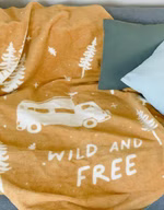 eine senfgelbe Kuscheldecke mit dem cremefarbenen Motiv von Bäumen, einem Campingbus und dem Schriftzug wild & free liegend auf einem Sofa mit grünen Kissen