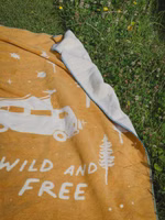 eine senfgelbe Kuscheldecke mit dem cremefarbenen Motiv von Bäumen, einem Campingbus und dem Schriftzug wild & free liegend auf einer grünen Wiese