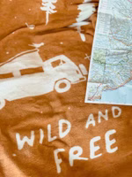eine senfgelbe Kuscheldecke mit dem cremefarbenen Motiv von Bäumen, einem Campingbus und dem Schriftzug wild & free liegend neben einer Landkarte
