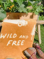 eine senfgelbe Kuscheldecke mit dem cremefarbenen Motiv von Bäumen, einem Campingbus und dem Schriftzug wild & free liegend im Garten mit Wanderschuhen