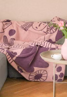 eine dunkel- und hell-lilafarbene Kuscheldecke mit dem Motiv von abstrakten Blumen und einer Gärtnerin liegend auf einer Couch im Wohnzimmer