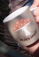 sonnig in der Hand haltend eine taupe-grün farbende Henkeltasse aus Porzellan mit dem dunkelbraunen Schriftzug Glückspilz und darüber ein Fliegenpilz mit pastellorangem Hut