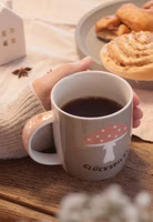 in der Hand haltend auf einem gedeckten Tisch seitlich eine taupe-grün farbende Henkeltasse aus Porzellan mit dem dunkelbraunen Schriftzug Glückspilz und darüber ein Fliegenpilz mit pastellorangem Hut sowie rosa weiß gepunktetem Henkel