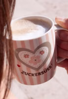in der Hand haltend und mit Milchschaum eine rosa cremeweiß gestreifte Henkeltasse aus Porzellan mit dem dunkelbraunen Schriftzug "Zuckersüss" und darüber ein großes Herz mit Blümchen