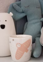 auf einem Nachttisch stehend neben Kuscheltieren ein kleiner cremeweiß farbender Porzellanbecher "Nachtaktiv" mit einer fliegenden rosa Eule sowie abstrakte Bäume und Sterne in taupe