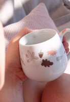 in der Hand haltend eine cremeweiße bauchige Henkeltasse aus Porzellan mit Blättern, Eicheln, Pilzen in herbstlichen Tönen braun, pastellorange und taupe