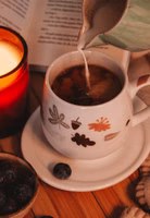 auf einem Tisch stehend und mit Tee befüllt eine cremeweiße bauchige Henkeltasse aus Porzellan mit Blättern, Eicheln, Pilzen in herbstlichen Tönen braun, pastellorange und taupe