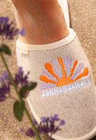 draußen ein sandfarbener Filzpantoffel hinter einem Zweig mit dem Stickmotiv einer aufgehenden Sonne in orange und lila sowie dem Schriftzug Sonnenschein in weiß