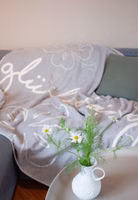 eine hellgraue Kuscheldecke mit cremefarbenen Glücksbringer Schriftzug sowie verschieden große Konturen von Kleeblättern liegend auf einem Sofa im Wohnzimmer