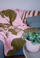 eine rosa Kuscheldecke mit dem olivgrünen, angeschnittenen Motiv eines Eukalyptus in einer gestreiften Vase liegend auf einem Sofa im Wohnzimmer