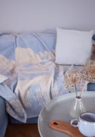 eine blau-graue Kuscheldecke mit einer cremefarbenen Berglandschaft und Almhütte im Vordergrund liegend auf einem Sofa im Wohnzimmer