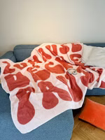 auf einem Sofa liegend eine rosa Kuscheldecke mit der dunkelrosa Aufschrift Zuhause Decke Pause