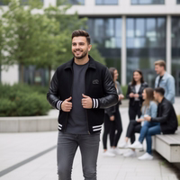 College Blouson Wool Leather Jacket A smiling man in a black college jacket with leather sleeves standing in front of a group of people sitting in a modern courtyard