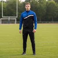 Model standing on sports field wearing STRIKER blue/black track suit with "German Wear" text. White stripe on chest and pants.