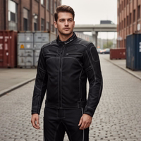 A man in a black motorcycle suit stands on a paved road in an industrial area surrounded by shipping containers