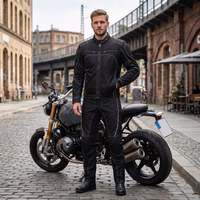 A man in a black motorcycle suit is standing next to a motorcycle on a paved road in front of a bridge