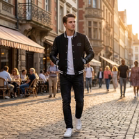 A man in a black college jacket with black leather sleeves and jeans walks down a paved sidewalk in the city